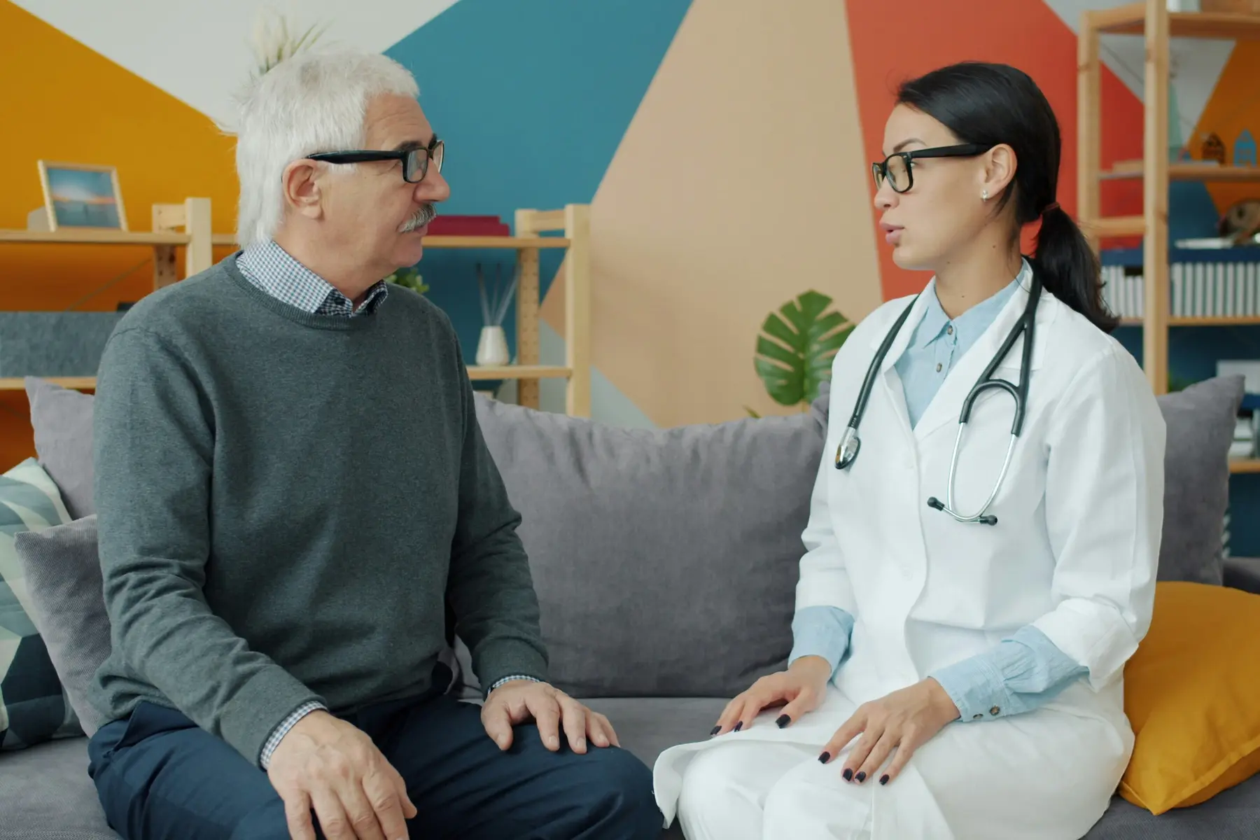 Home health professional with a stethoscope talking with a senior man on his living-room sofa
